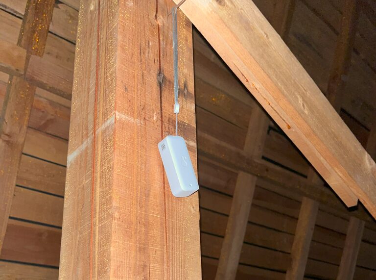 小屋裏の木材の柱に、白いSwitchBot防水温湿度計を吊り下げて設置した写真。天井点検口から安全に設置し、小屋裏の環境を遠隔でモニタリングする様子を示している。